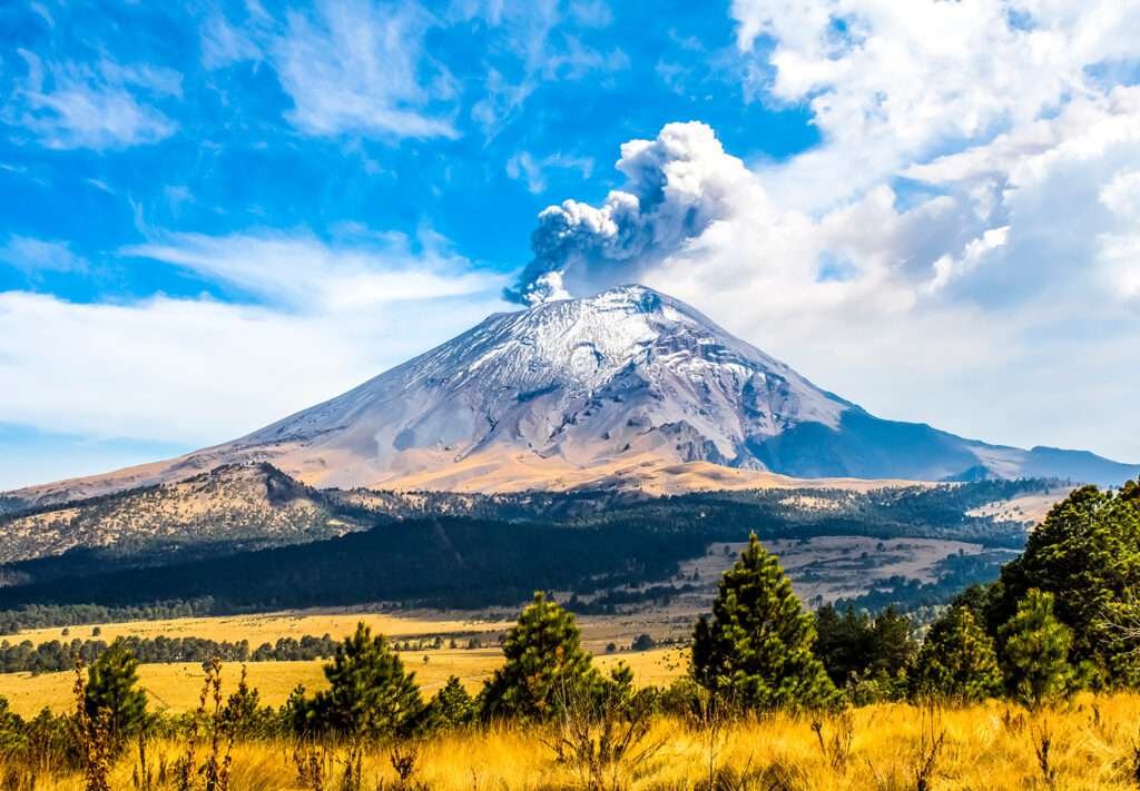 Der Popocatépetl, einer der drei höchsten Berge Mexikos, mit Aschewolke vor blauem Himmel und Vulkanlandschaft im Vordergrund
