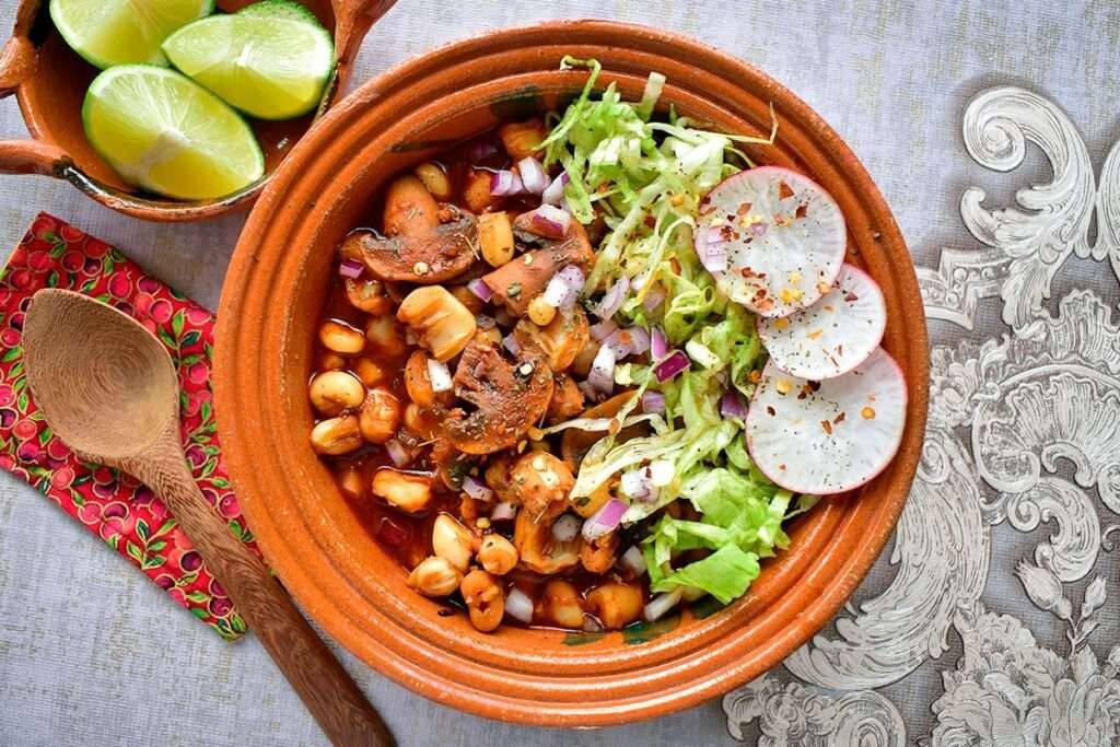 Mexikanische Pozole Rojo in Tonschale mit Pilzen, Mais, Radieschen, Zwiebeln und Eisbergsalat